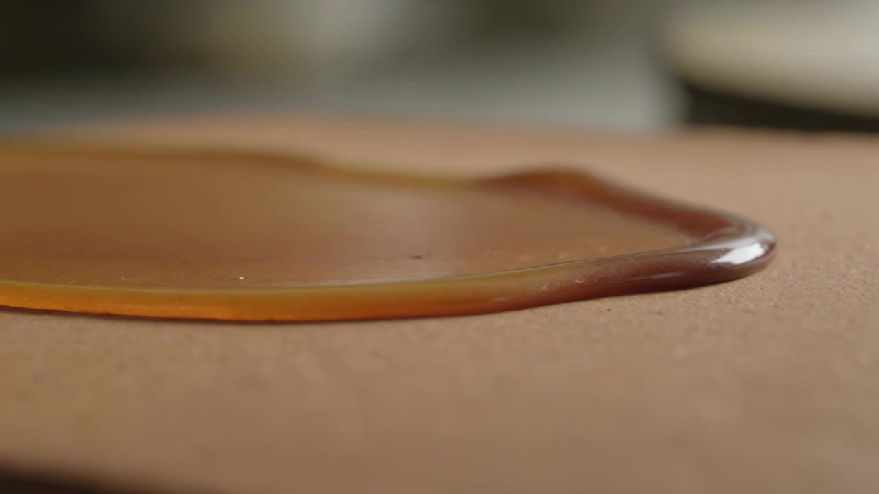 Charco ámbar nivelándose y extendiéndose sobre una tabla marrón, impulsado por la tensión superficial, con utensilios de cocina borrosos.
