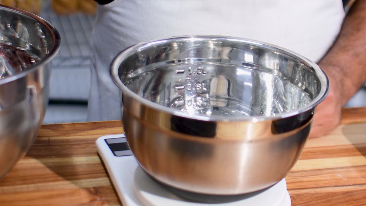 Närbild på händer som mäter ingredienser på en elektronisk våg i en metallskål i ett rustikt träkök, vilket visar noggrann bakförberedelse