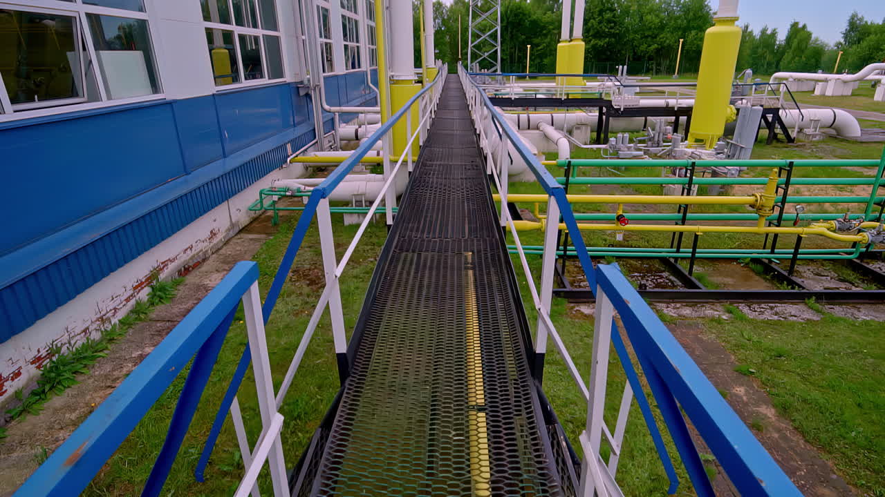 Elevated metal walkway running through industrial gas plant with visible pipelines and tanks