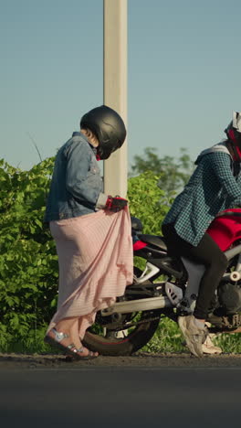 dos mujeres con cascos, una sentada en una bicicleta deportiva roja preparándose para moverse, mientras que la otra dama levanta su bata mientras sube a la bicicleta