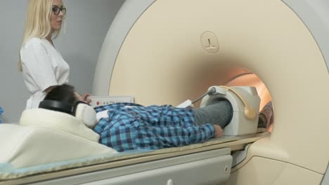 woman doctor makes knee-joint mri scanning. young man patient on automatic mri table enters into a closed-type magnetic resonance imaging machine using noise isolation headphones. modern equipment, coil on the patient's knee.