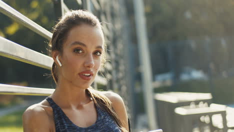 Portrait-Of-A-Cheerful-Sporty-Woman-With-Airpods-Smiling-At-The-Camera-After-Workout-At-Outdoor-Court-On-Sunny-Day