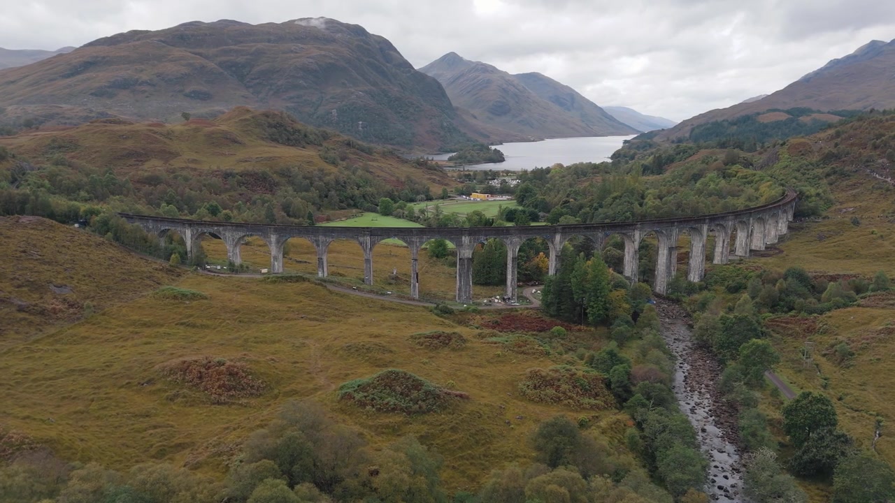 Vuelo circular aéreo alrededor del Viaducto de Glenfinnan y el Lago Shiel, Escocia, con vistas panorámicas del viaducto y el paisaje circundante, vías de tren icónicas con soportes arqueados.