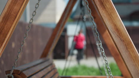 close up of wooden swing structure with metal chains under sunlight, background blurred with people walking near fence and building, creating peaceful outdoor atmosphere