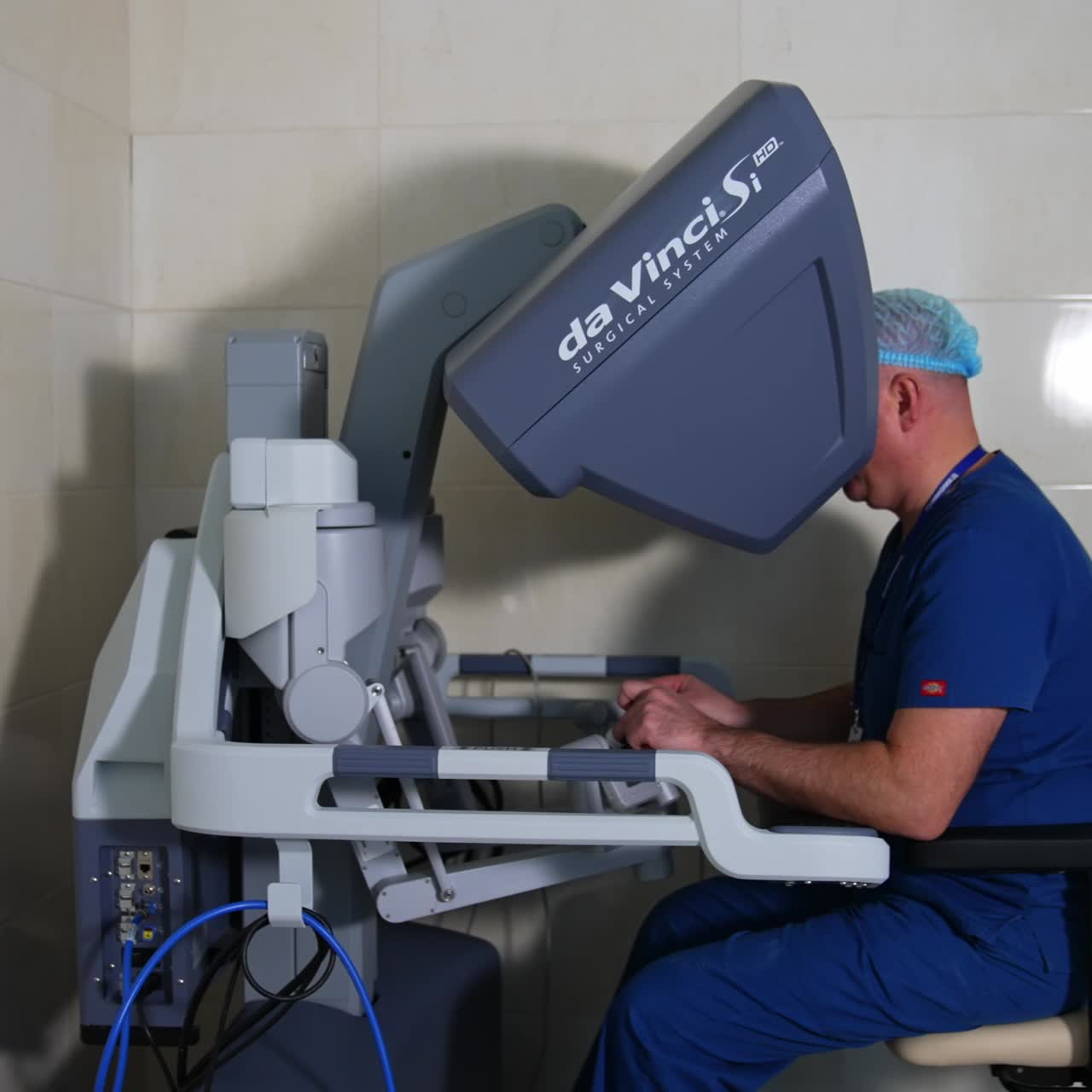 Adult male surgeon in cap and uniform sits at the control unit of Da Vinci surgical system. Doctor looks to image processor and moves manipulators