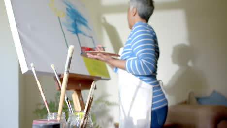 mujer biracial mayor con delantal y pintando en un lienzo grande en casa, en cámara lenta