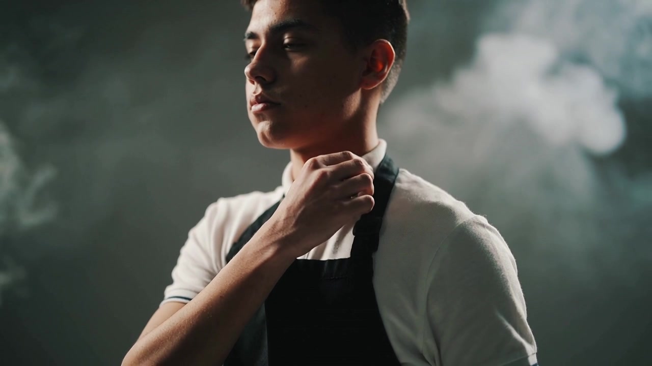 Uomo che tocca la polo bianca e il grembiule nero regolando la cinghia, in preparazione per il servizio nella nebbia di studio