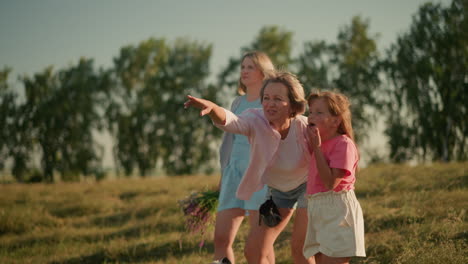 family standing on hilly farmland as mother points out something interesting in distance to her daughter while another woman stands in background holding flowers