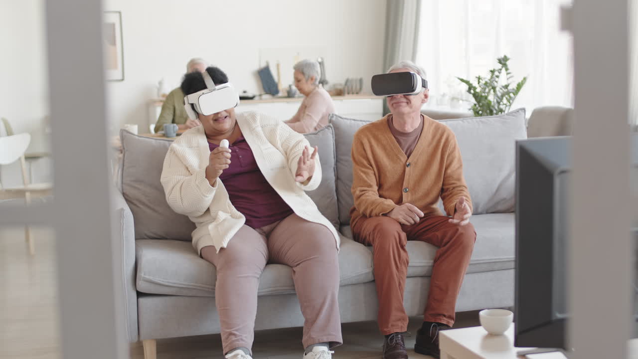 Full shot of amused man and woman wearing virtual reality headsets, sitting on couch in living room, moving heads around, laughing