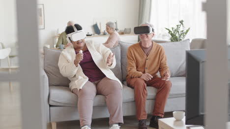 full shot of amused man and woman wearing virtual reality headsets, sitting on couch in living room, moving heads around, laughing