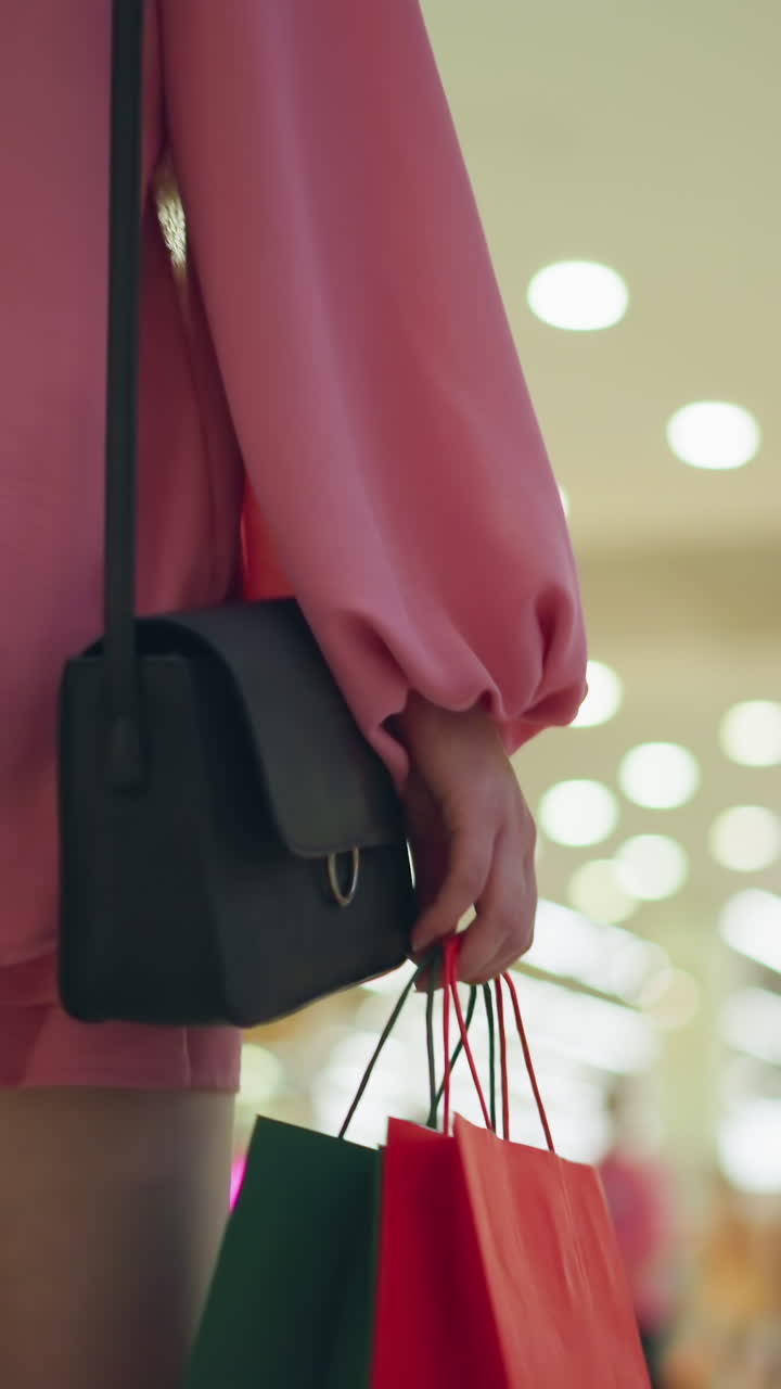 핑크 을 입은 여성의 뒷면, 밝은 불빛으로 조명된 쇼핑몰을 는 동안 양손에 다채로운 쇼핑 가방을 들고, 다른 쇼핑객의 흐릿한 시야와 배경의 천장 불빛