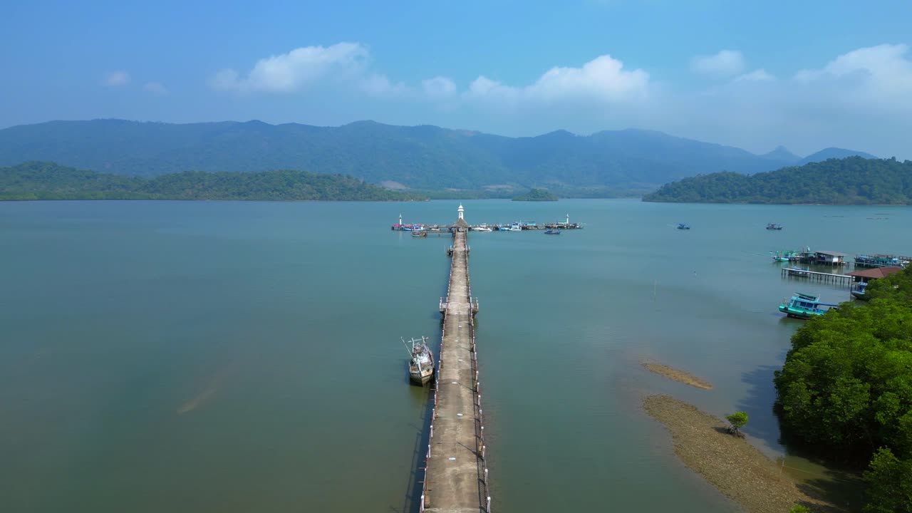 Long Walkway pier with a lighthouse reaching into blue ocean with mountains under clear sunny sky. Gorgeous aerial view overflight flyover drone