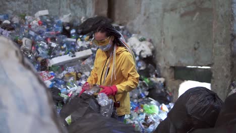 Stylish-woman-in-yellow-jacket,-protective-glasses-and-gloves-sorting-plastic-bottles-from-black-bags.-Huge-pile-of-used-plastic-bottles-on-background