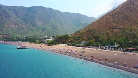 Luftdrohne,-Die-An-Einem-Heißen-Sommertag-In-Der-Türkei-Die-Blaue-Mittelmeerküste-Des-Adrasan-strandes-Voller-Touristen-Und-Einer-Trockenen-Berglandschaft-Schwelgt