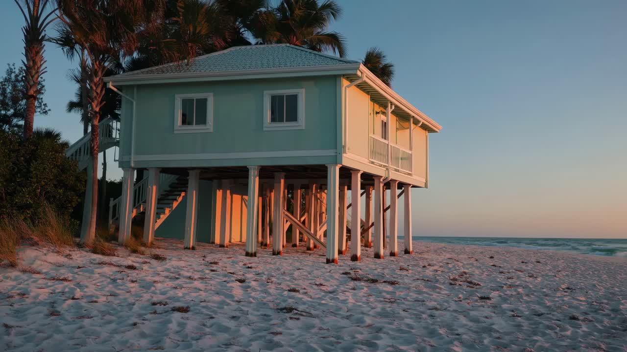 Wooden stilt house perched over sandy tropical shoreline, silhouetted against golden sunset with swaying palm trees creating serene island atmosphere