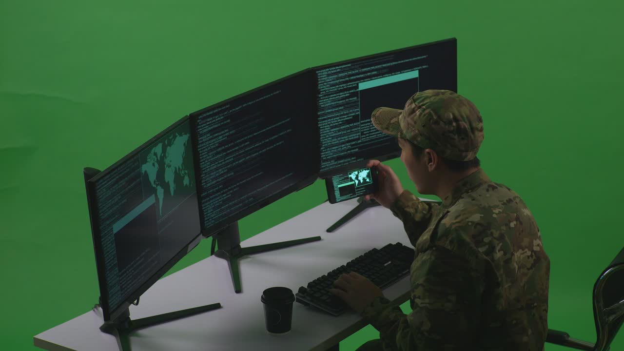 Side View Of Happy Asian Man Soldier Developer Looking At Code On Mobile Phone While Write Code With Multiple Computer Screens In Green Screen Studio