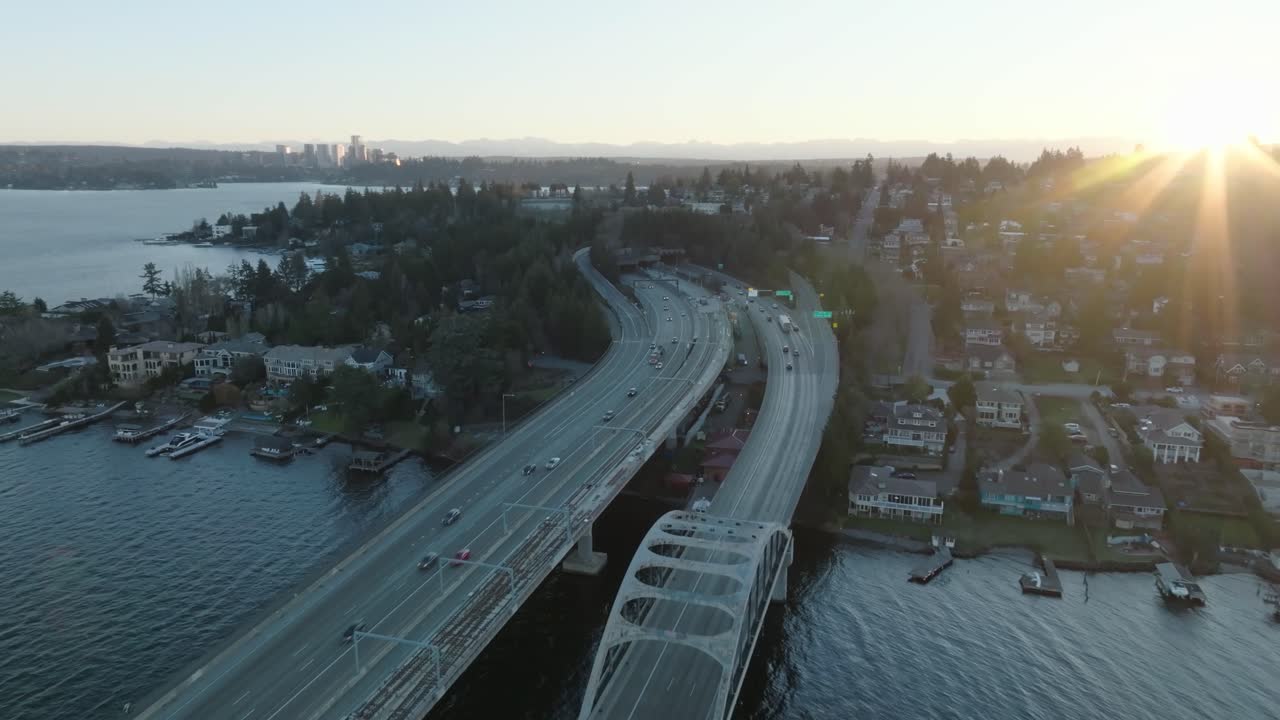 Drone flight captures cars approaching the floating bridge to Seattle’s serene island. The tranquil blue water and distant sunrise create a captivating scene.