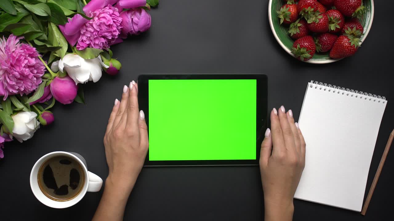 Woman working on tablet computer device with green screen. Beautiful white and pink peonies flowers, strawberries, coffe and notebook on the black background. Table top view. Chroma key