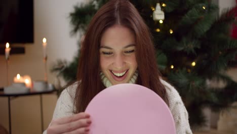 A-happy-brunette-girl-opens-a-pink-round-box-of-her-gift-and-looks-at-what's-inside-near-the-Christmas-Tree-in-a-cozy-room-decorated-in-Christmas-style-in-the-evening