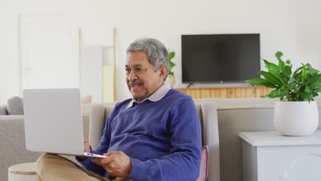 Video-De-Un-Hombre-Birracial-Senior-Feliz-Usando-Una-Computadora-Portátil-Sentado-En-La-Sala-De-Estar,-Sonriendo