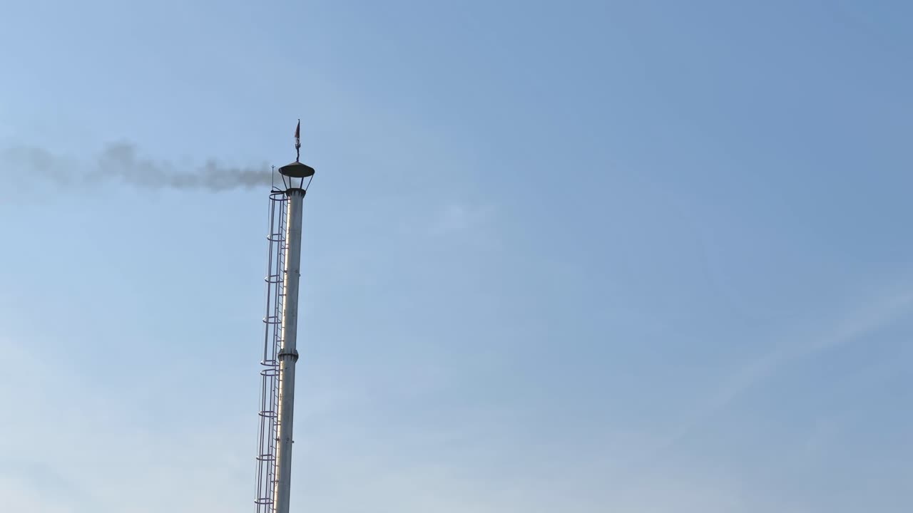 Tall industrial chimney releases a thin stream of smoke into a clear blue sky, showing its laddered design and upper vent structure in a simple static frame
