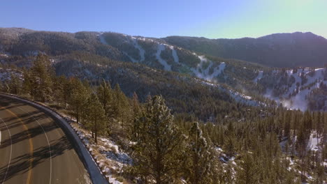 Boom-down-towards-road-winding-through-mountains-in-Lake-Tahoe,-Nevada-on-a-beautiful-day-with-Douglas-Firs-in-the-foreground-and-mountain-peaks-on-the-horizon