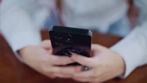 blurred view of lady hand with black polished nails engaging with smartphone placed on wooden table, surrounding blurred background with green and pink unclear items