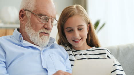 Close-Up-Of-The-Senior-Grey-Haired-Grandpa-In-Glasses-Demonstrating-Old-Photos-To-His-Pretty-Granddaughter-While-They-Sitting-On-The-Couch-At-Home