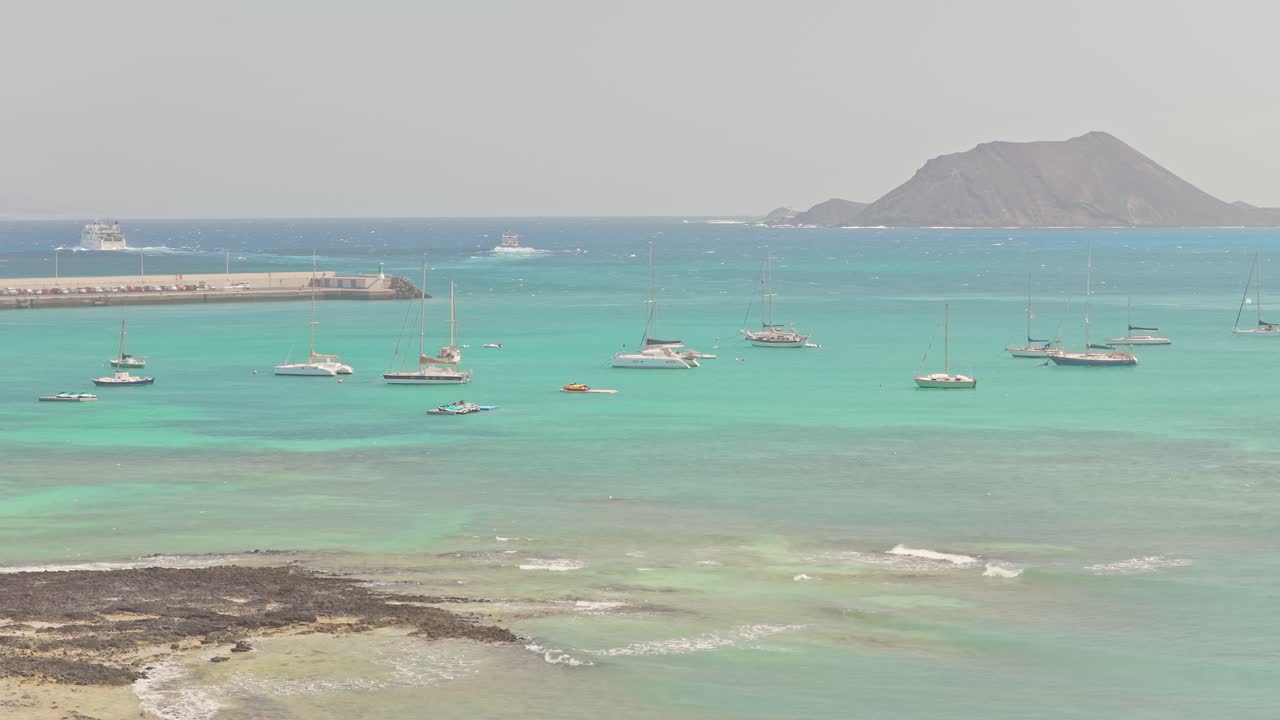 Midday drone pull-out over Playa del Medio Beach Area in Fuerteventura, Canary Islands, Spain, revealing turquoise waters, anchored sailboats, and a mountainous island backdrop