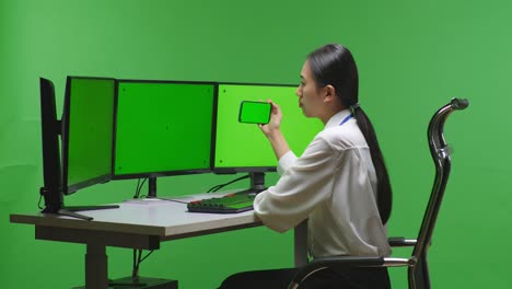 woman using smartphone on green screen in front of multiple monitors