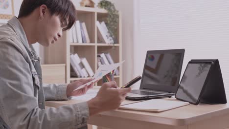 asian designer man uses his smartphone to communicate. work on projects and papers. and work from home on a desk with a laptop.