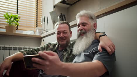 a happy elderly man with gray hair and a lush gray beard in a gray t-shirt sits on the floor with his brunette boyfriend with stubble and a green checkered shirt and looks at their photos on a smartphone in the kitchen during the day