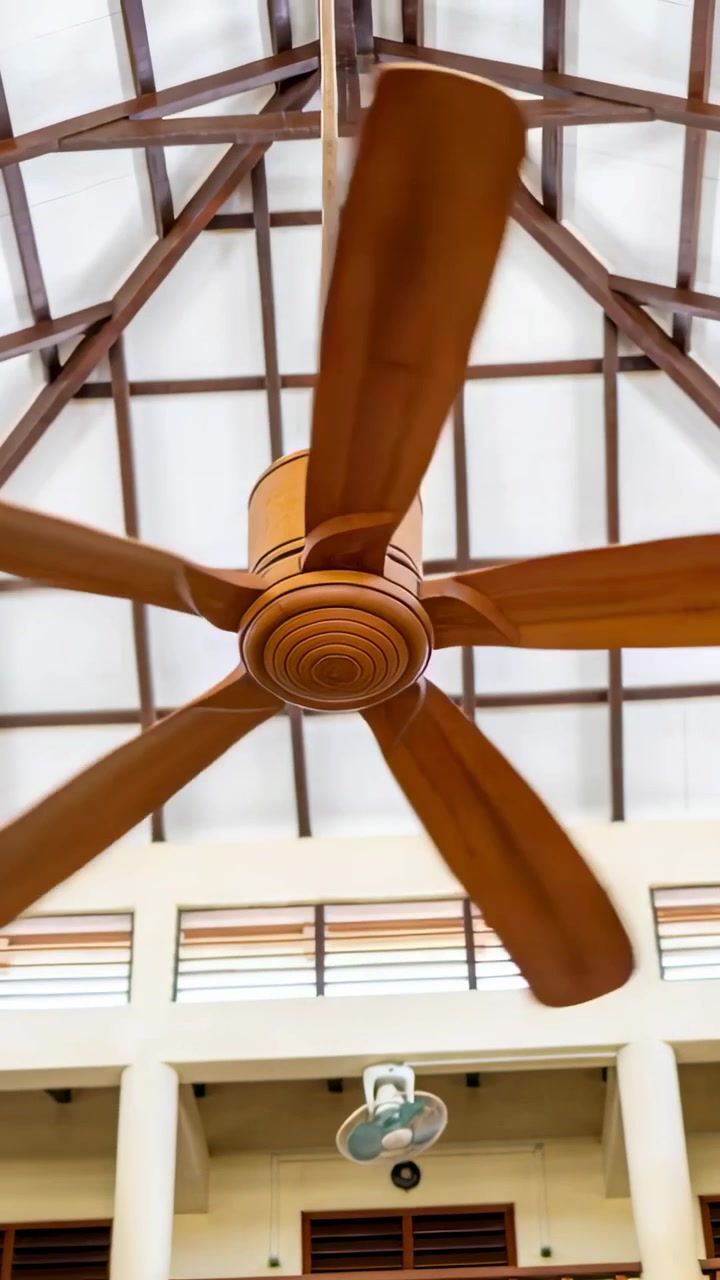 Vertical video: Large ceiling fan powering up, spinning blades sweeping frame amid trusses for HVAC