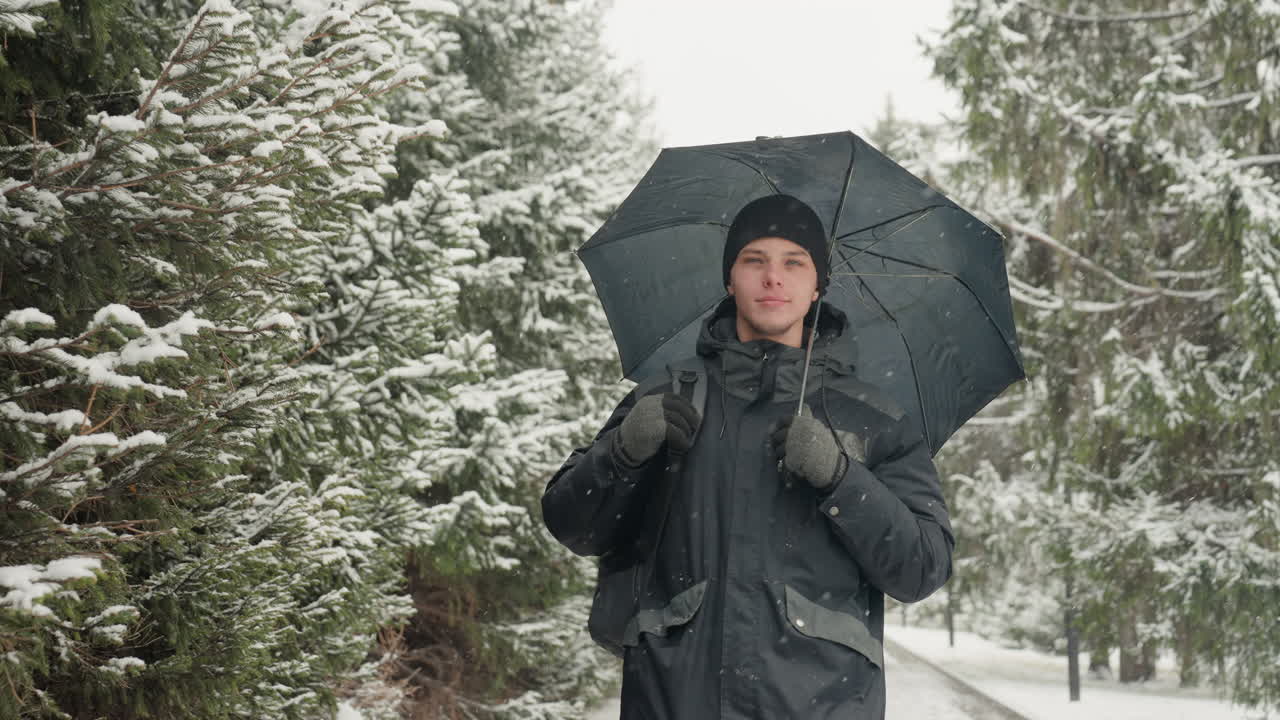Man holding umbrella in snowy forest clearing, tall pine and evergreen trees laden with snow, visible breath in cold air, paused gaze and cinematic portrait vibe, rugged coat and mittens