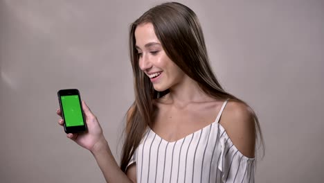 young excited girl is showing green screen of smartphone, wow, watching at camera, smiling, communication concept, grey background