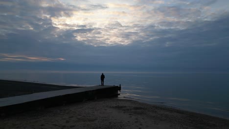 Man-walking-to-jetty-end-at-overcast-dusk-with-coastline-reveal-of-Fleetwood-Beach-Lancashire-UK