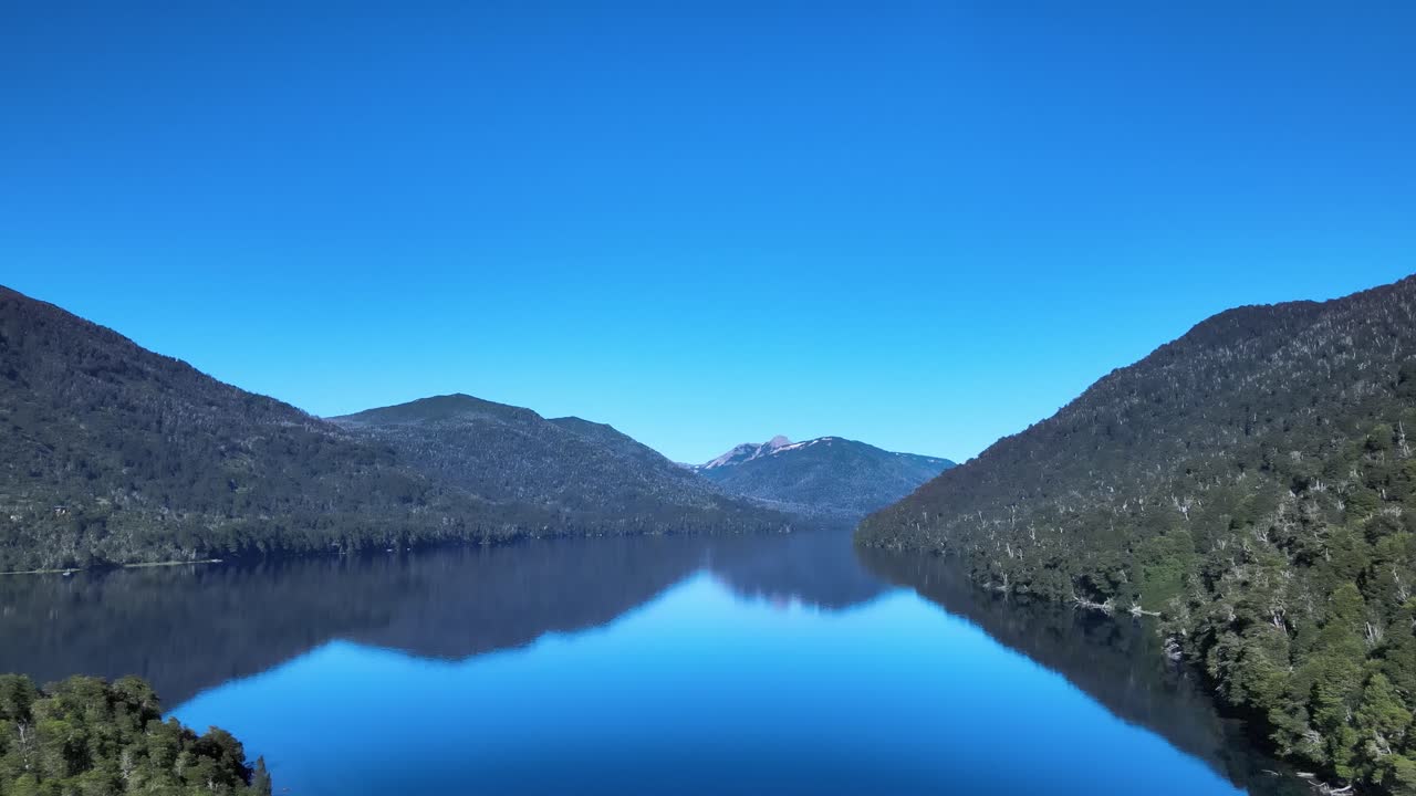 A blue sky day, through the forest, penetrating the mirror of Lake Hermoso. Patagonia, Argentina. Aerial footage.