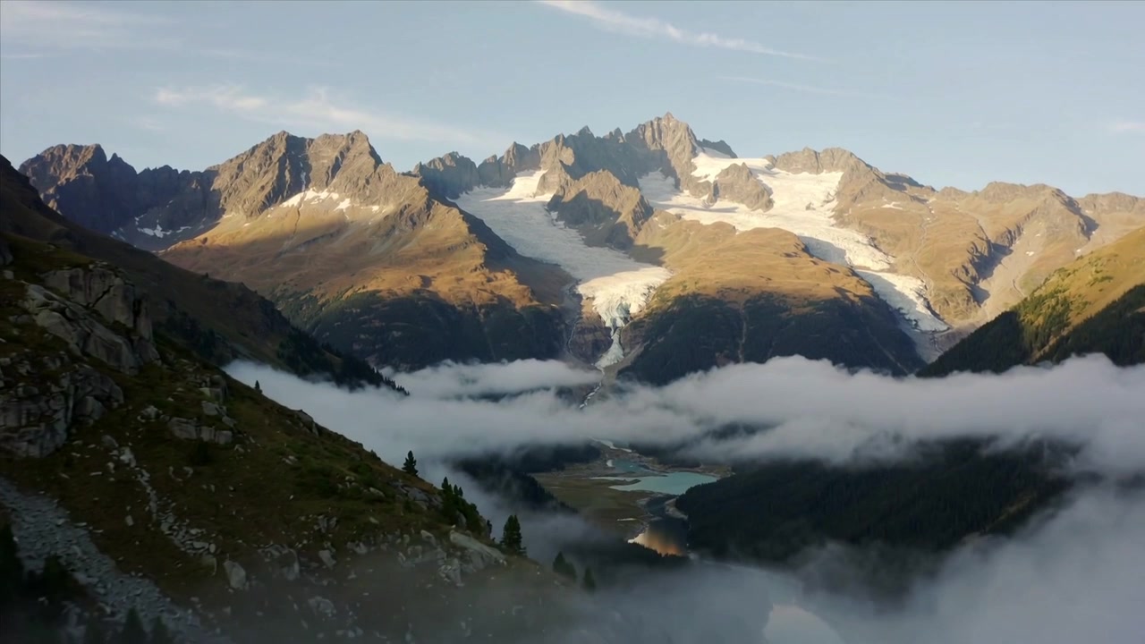 Une caméra panoramique révèle une vallée alpine, la lumière du soleil dissipant le brouillard et dévoilant une rivière bleue et des glaciers.