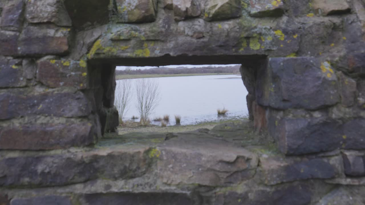 A slow camera move through a rectangular opening in a brick wall reveals a lake at Rainton Meadows Nature Reserve. Sand, soil, reeds and bushes lead to calm water, with trees and cloudy skies beyond