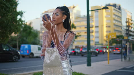 brown skin lady looking hand mirror at evening street. woman applying makeup