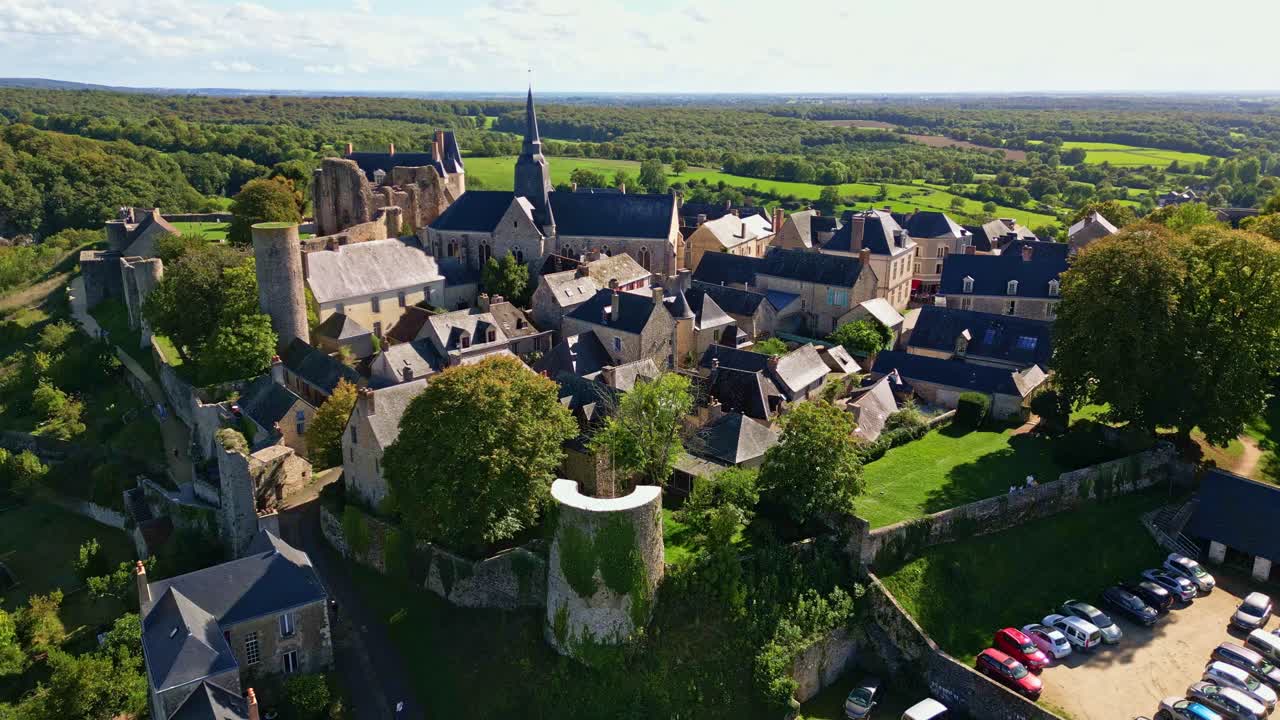 Drone ascending and tilt-up above the medieval city of Sainte-Suzanne in Mayenne, showing the castle, village houses, and nearby parking area under a sunny sky