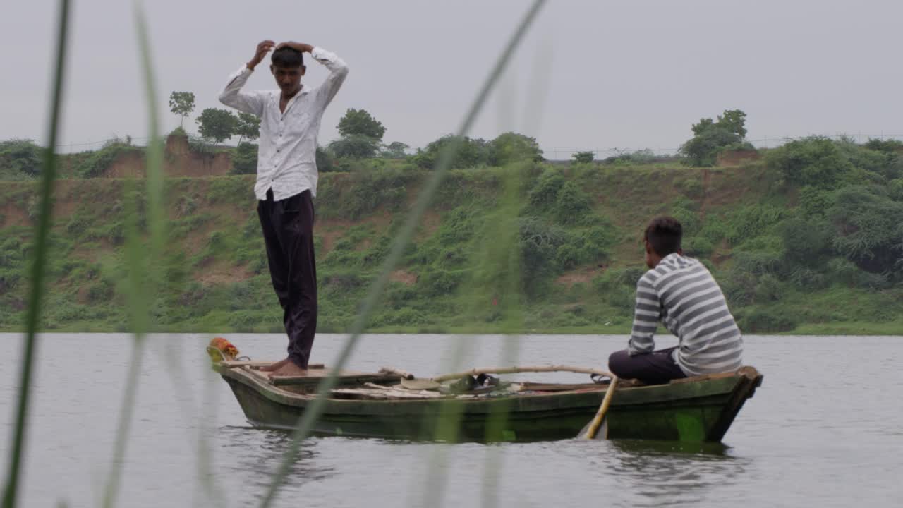 una cámara lenta de mano larga toma de primer plano de dos hombres indios en un pequeño bote de madera, uno sentado y remando y el otro de pie, con el primer plano de hierba silvestre en el frente mientras se mueven en el agua del río