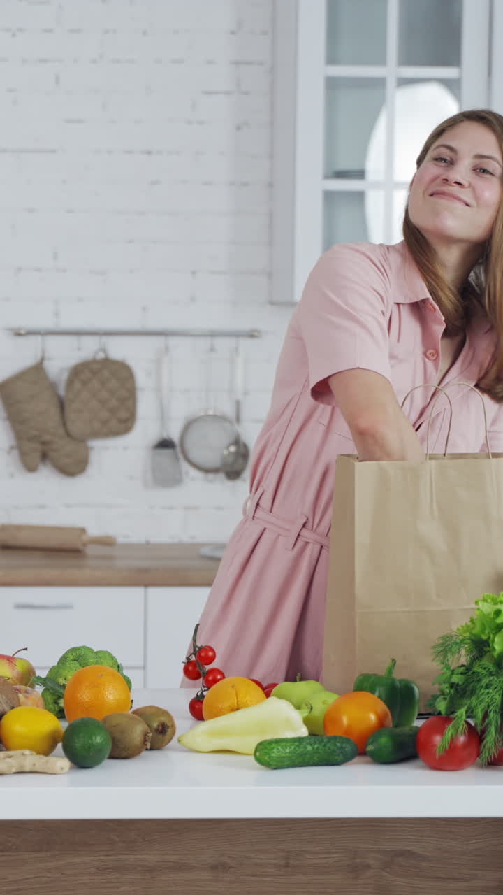 Joyful woman taking out fresh fruit from paper bag in the kitchen. Organic fruit and vegetableson the table and beautiful female indoors. Healthy lifestyle. Vertical video