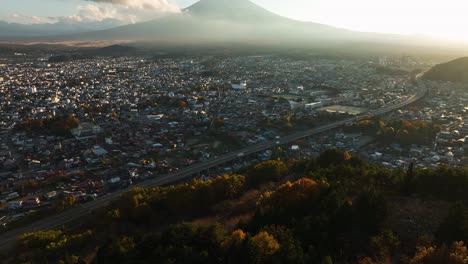 Vista-Aérea-Sobre-El-Bosque-De-Colores-Otoñales,-Hacia-Una-Ciudad-Y-El-Monte-Fuji,-Hora-Dorada-En-Japón