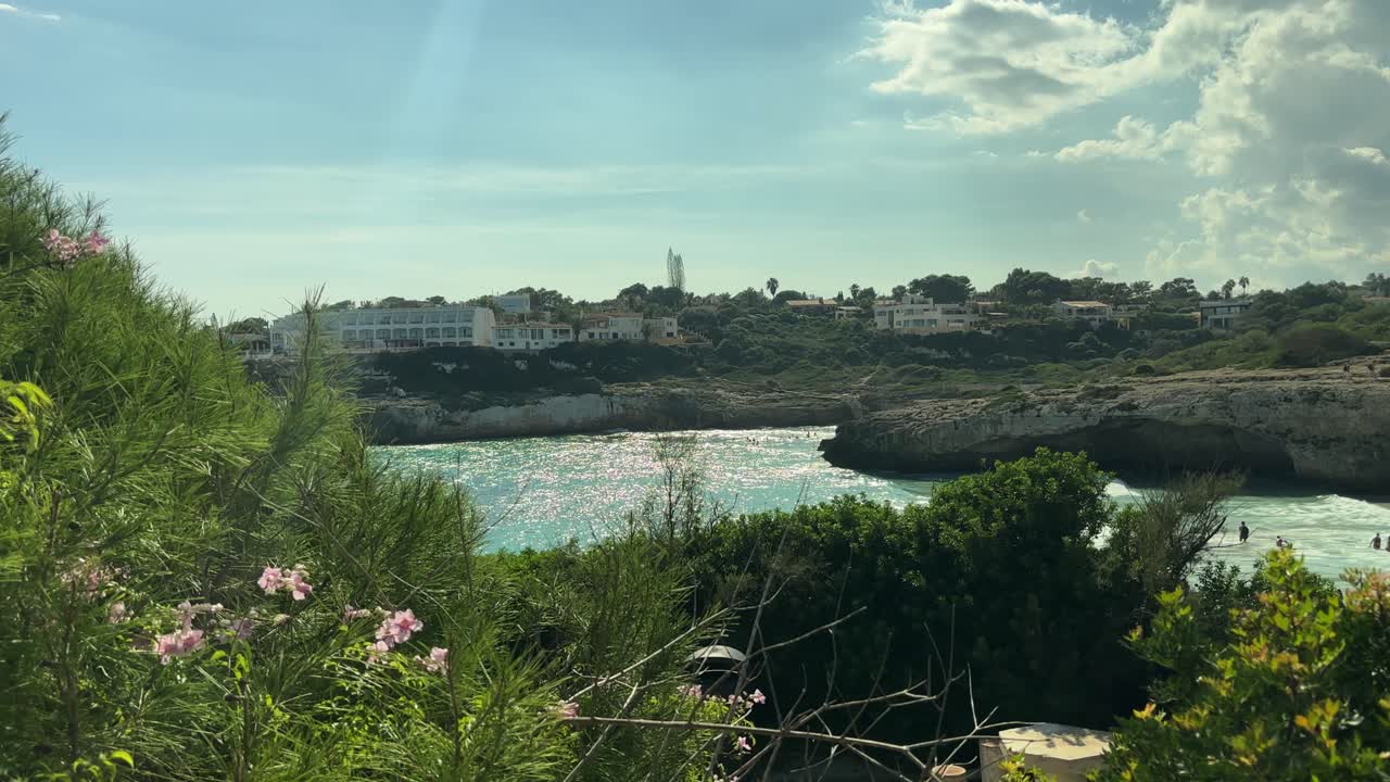 꽃과 초목으로 둘러싸여 있고 청록색 바다가 보이는 스페인 마요르카 해안의 바위 덮인 작은 만이 햇빛에 비치는 풍경이에요.