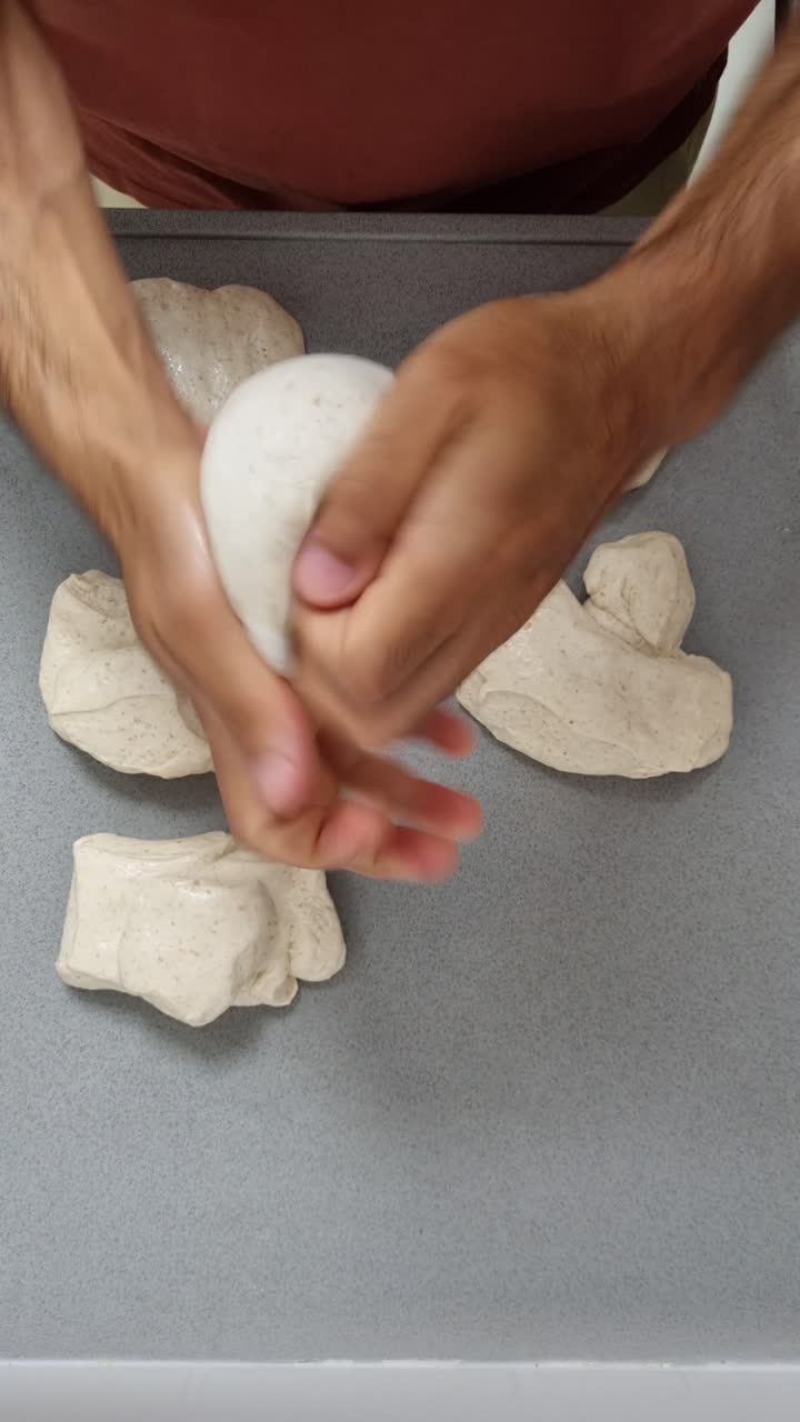 Transformação da massa de pizza fermentada a frio em esferas perfeitas através de movimentos profissionais de esticar e dobrar com as mãos, capturado em vídeo vertical aéreo
