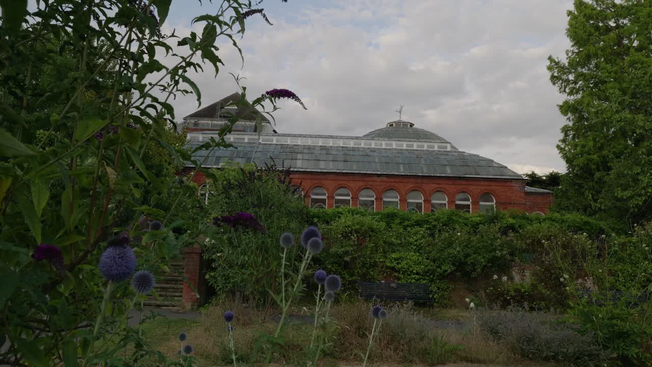 4K pan across Avery Hill Park revealing the historic Victorian Winter Garden with red brick facade, lush flowers, and Edwardian estate grounds in south east London