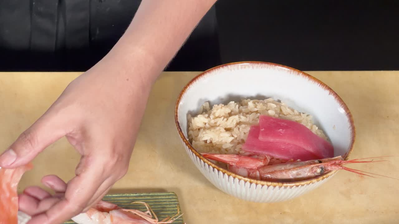 Overhead view of chef arranging sashimi and shrimp on rice bowl under soft studio lighting