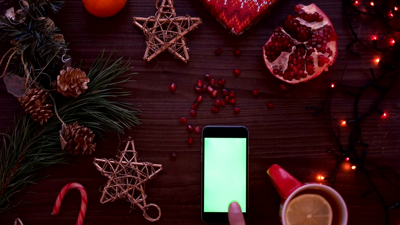vista superior hombre mano usando teléfono inteligente con pantalla verde. dedo desplazamiento de páginas en pantalla táctil. detalles de navidad en fondo de mesa de madera. llave de croma