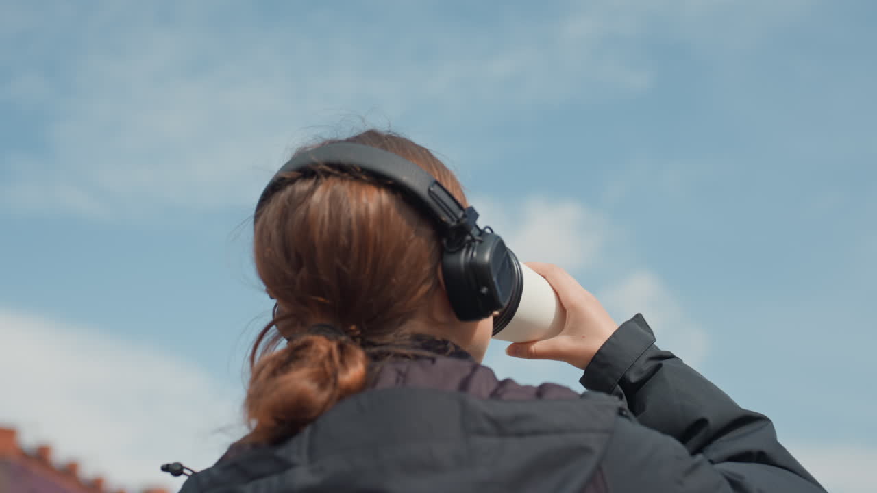 Persona dai capelli rossi che ascolta musica all'aperto tra gli edifici della città, indossa una giacca nera e uno zaino, cuffie grandi, guarda il cielo blu con nuvole soffici, stato d'animo riflessivo e calmo